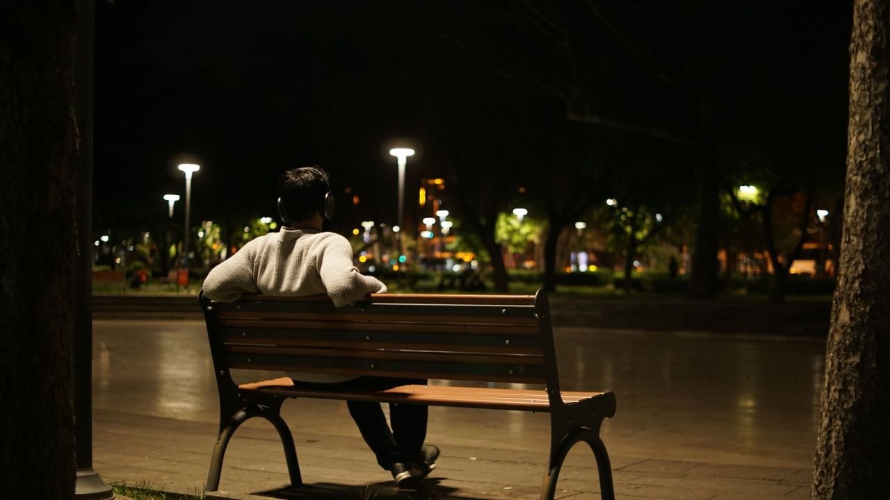 A man sitting alone on a park bench under street lamps at night