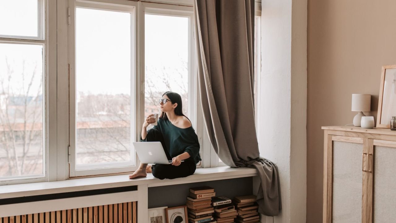 A woman sitting on a windowsill with a hot drink, looking out
